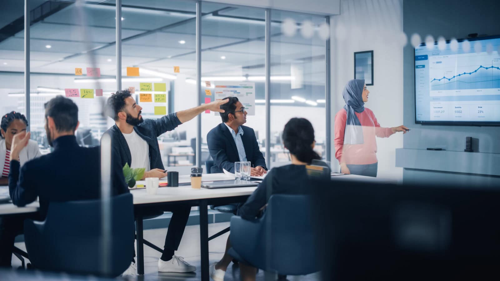 Multi Ethnic Office Conference Room. Muslim Female Ceo Wearing Hijab Does Presentation For Group Of Managers Talk, Use Tv Infographics, Statistics. Businesspeople Brainstorm Ecommerce Growth Strategy